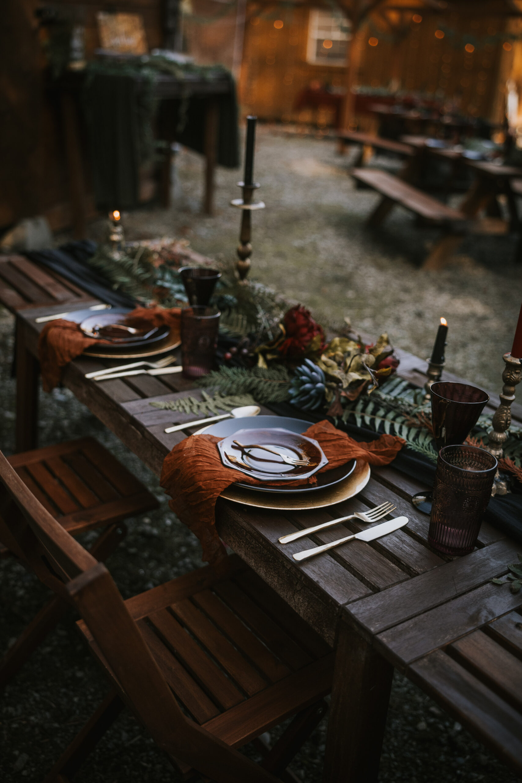 Long dinner table with rust linen napkins, brass candlesticks and fern runners at dusk