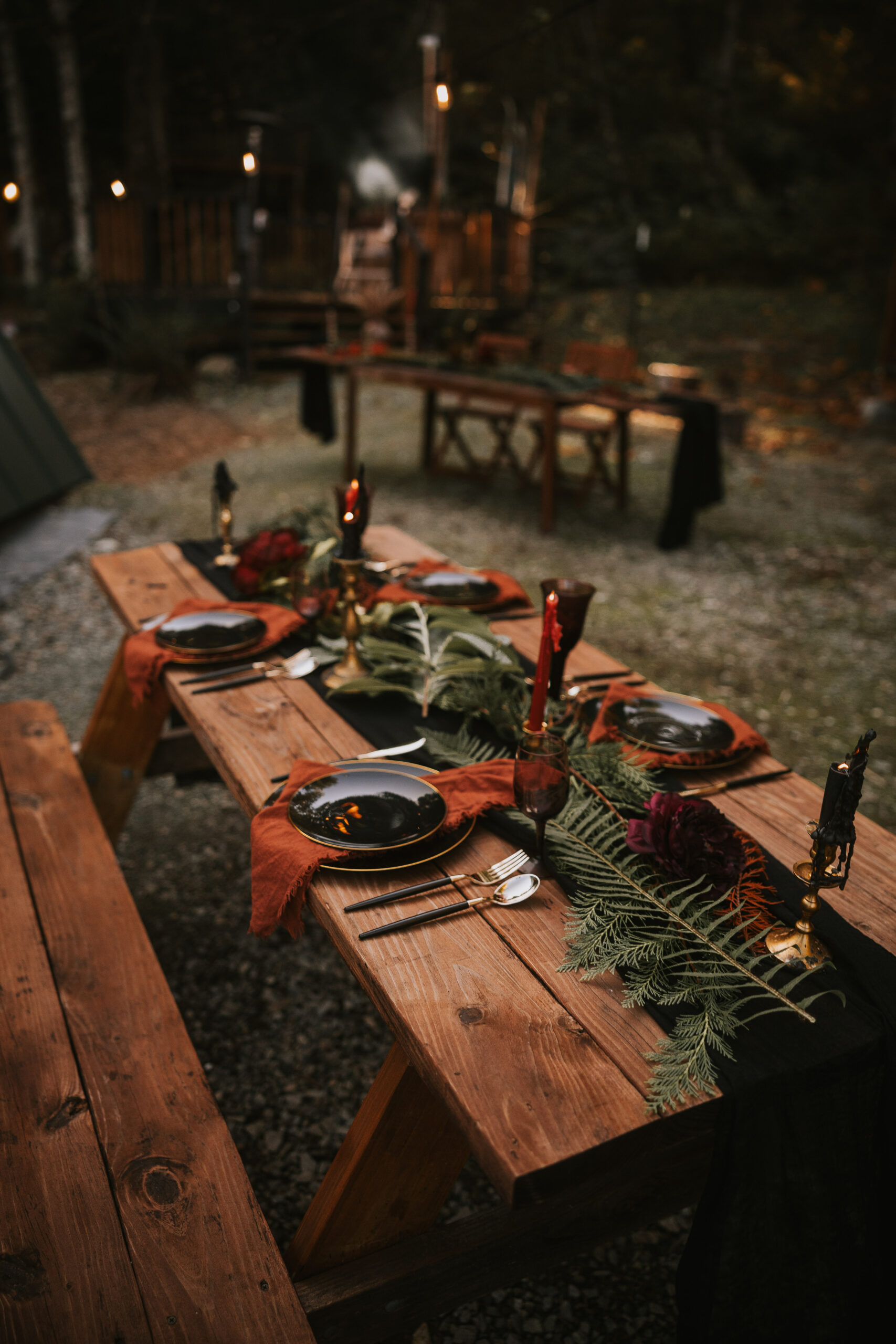 Picnic tables laid with black plates, rust napkins and fern garlands, the cabin lit behind