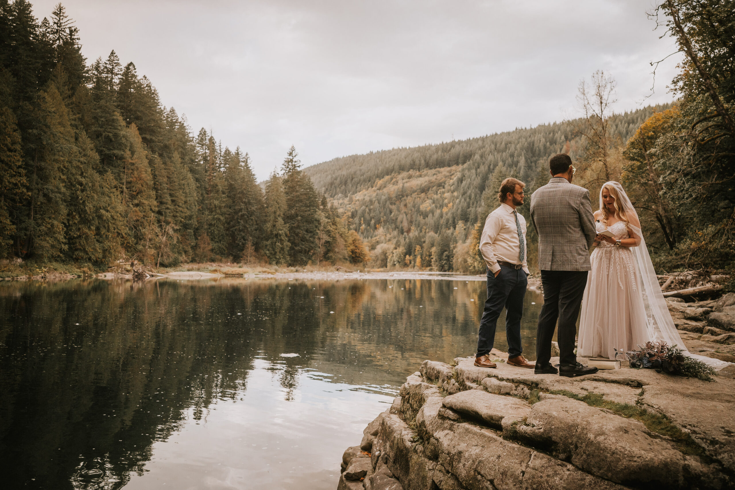 Wide view of the lakeside ceremony on the rocks, forested hills rising on the far shore
