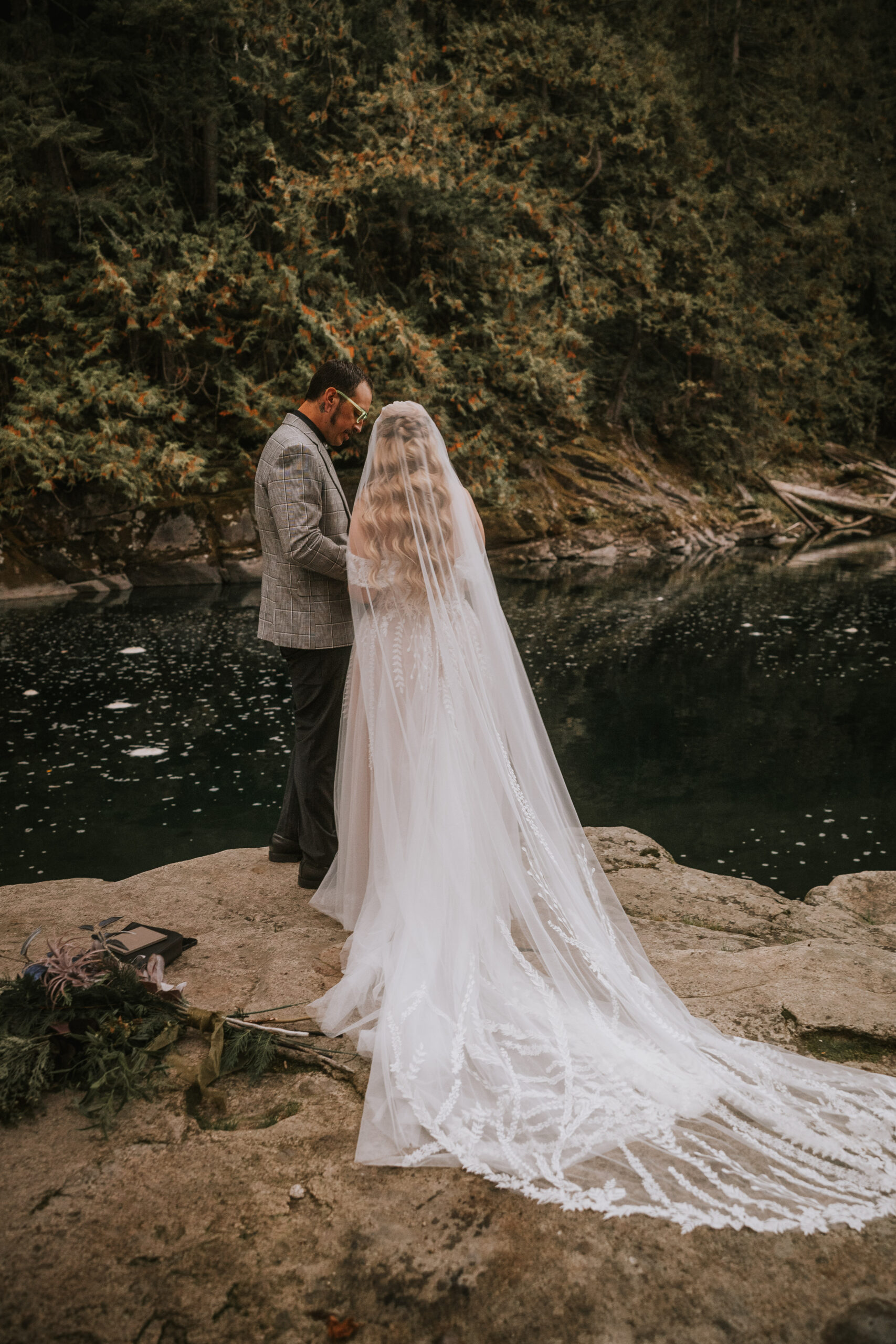 Justine and Zack at the river's edge, her cathedral veil spread across the rock behind them