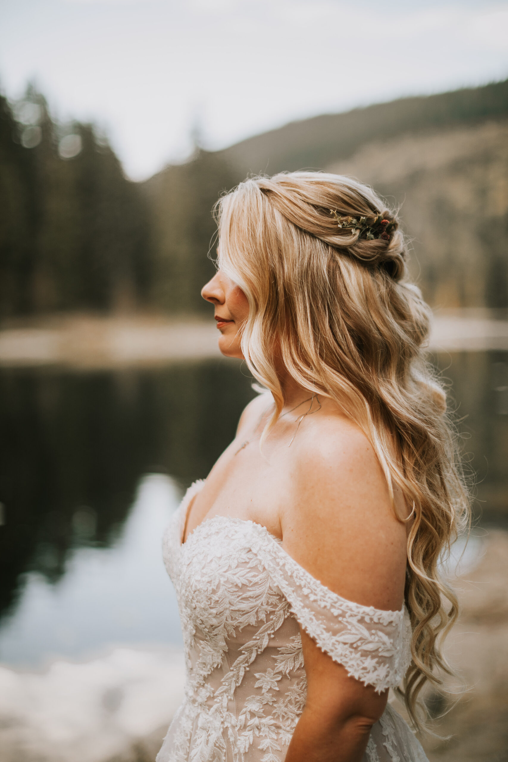 Justine in profile beside the lake in an off-shoulder lace gown, sprigs pinned into her braided hair