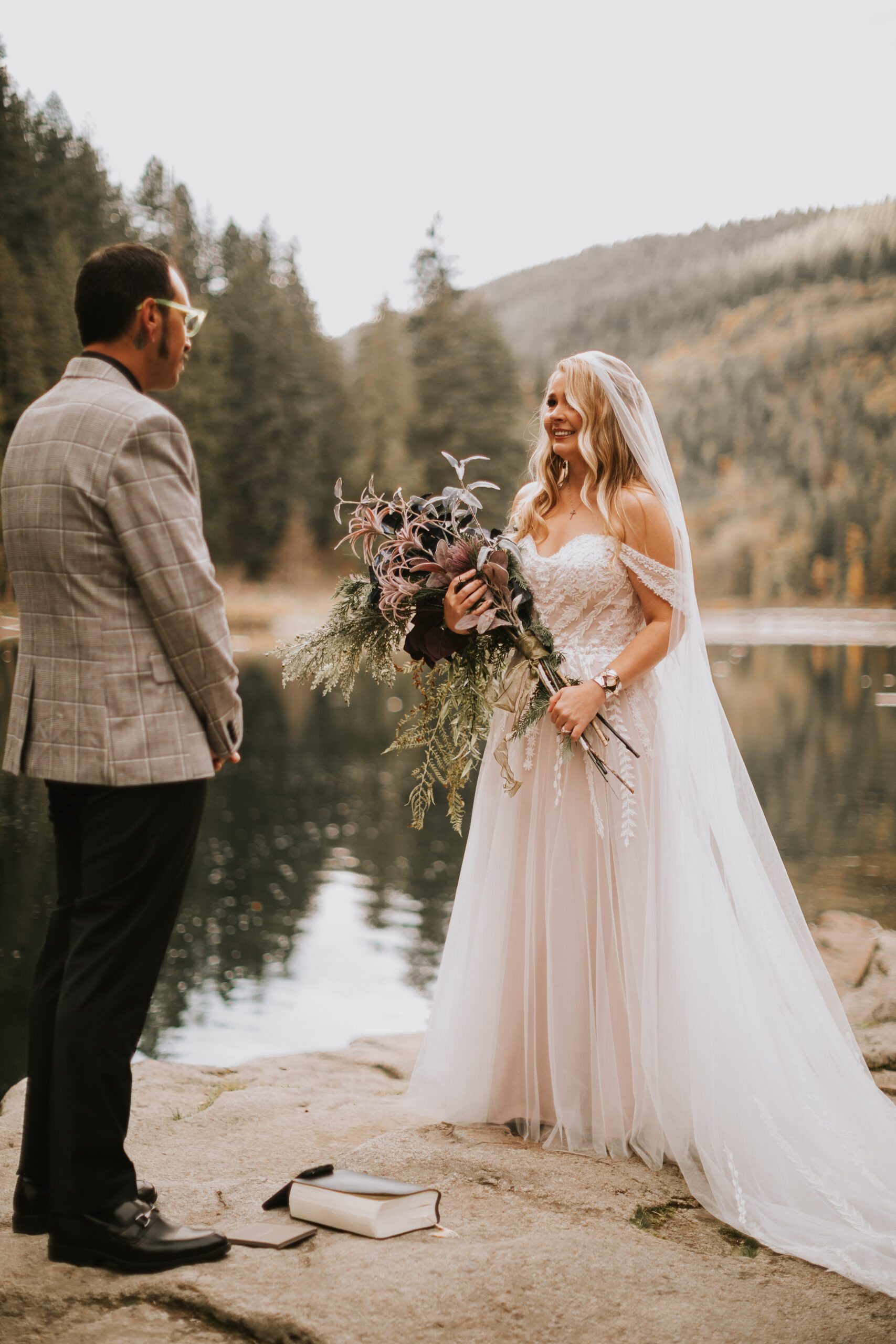 Justine and Zack facing each other at the water's edge, her dried bouquet in hand and their vow books on the rock below