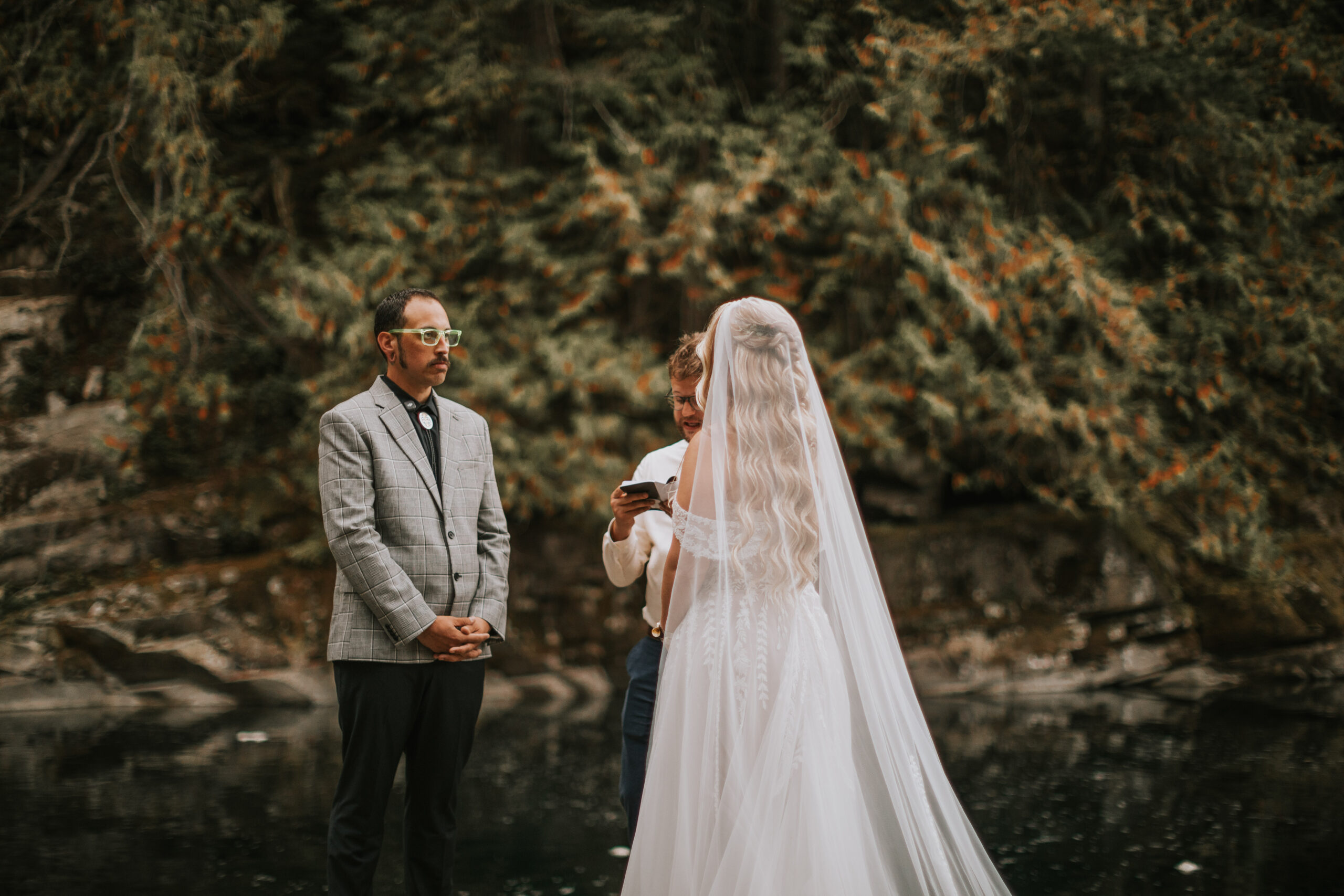 Justine from behind in her cathedral veil, facing the officiant with the forest and lake beyond