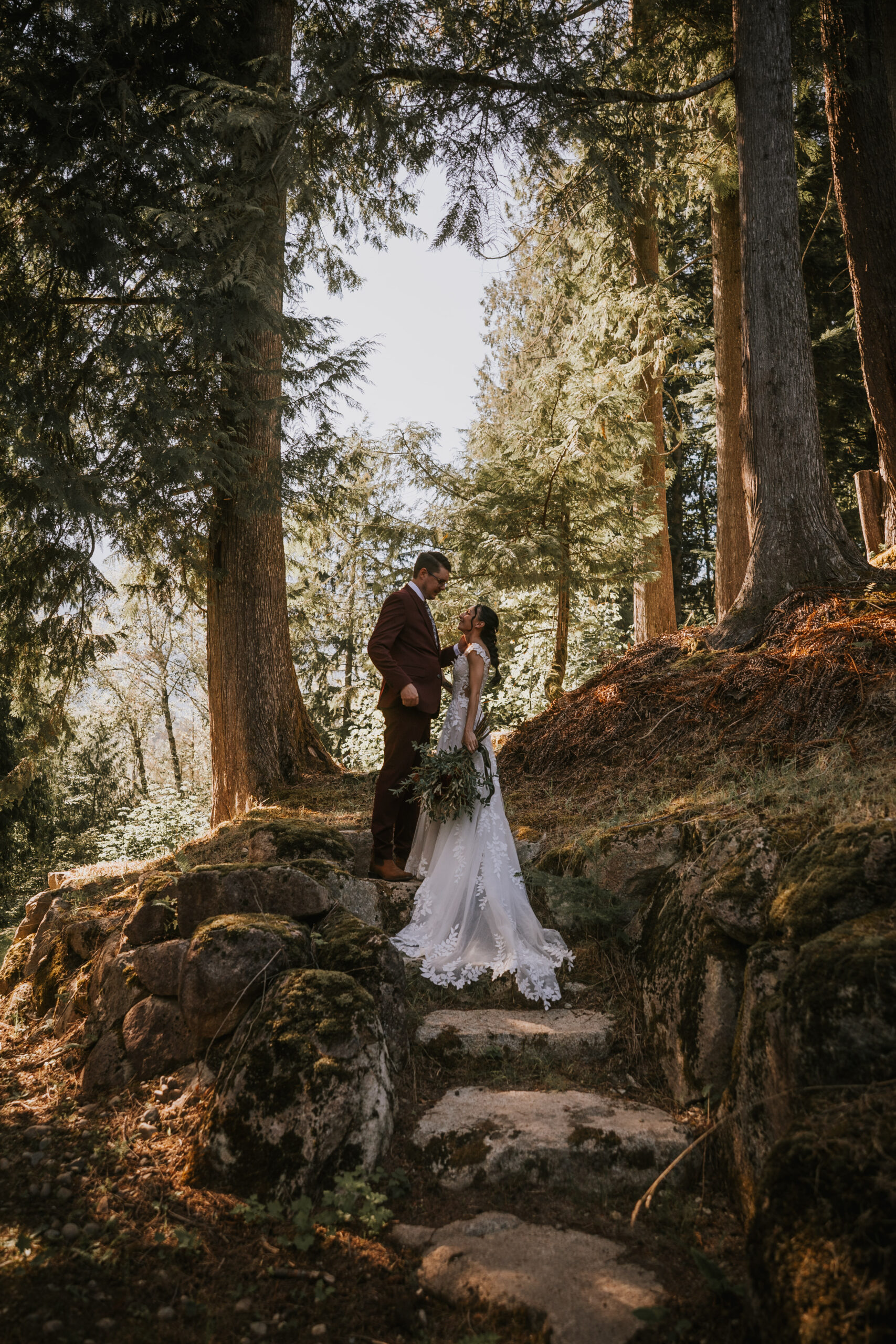 Brandon and Morgan on the mossy stone steps in the forest, greenery bouquet in her hand
