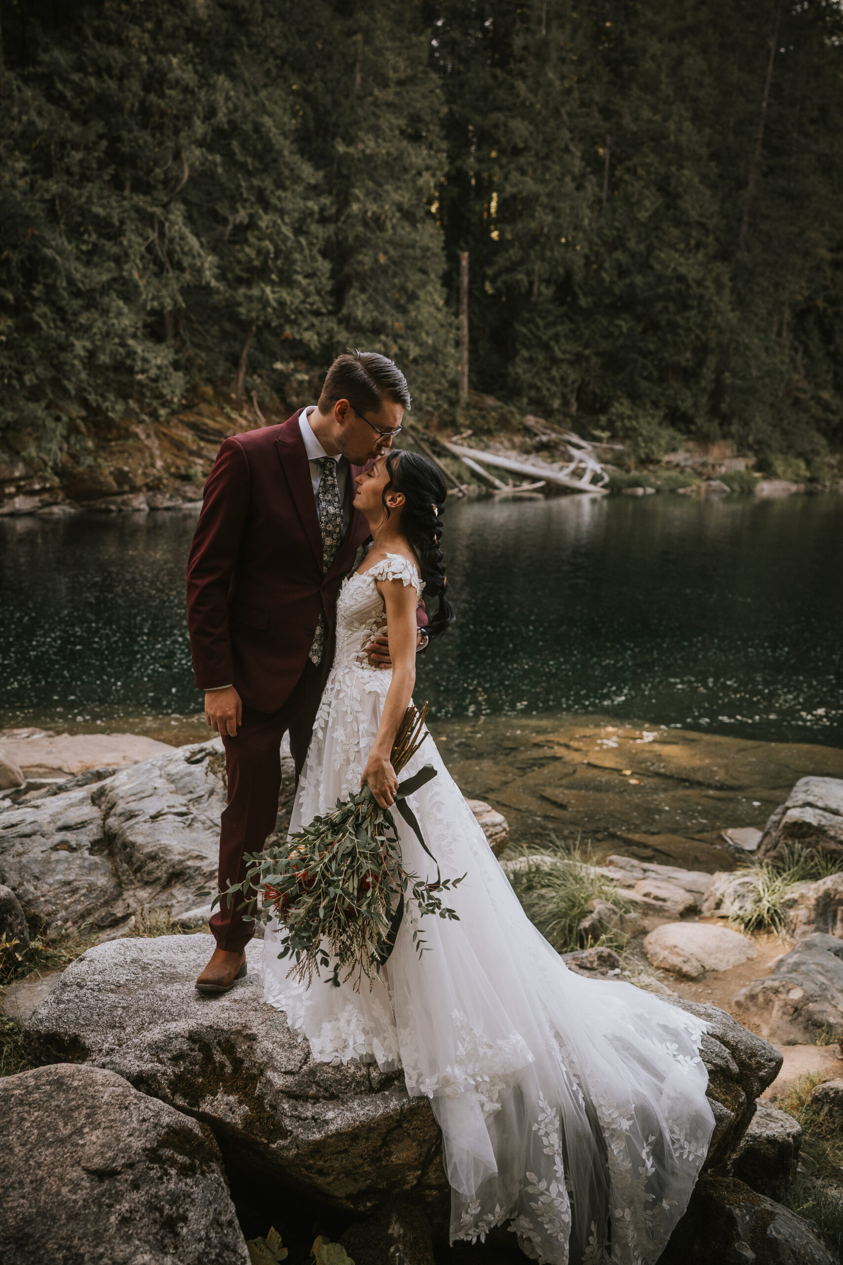 The couple kissing on river rocks, her long trailing bouquet of olive and eucalyptus in view