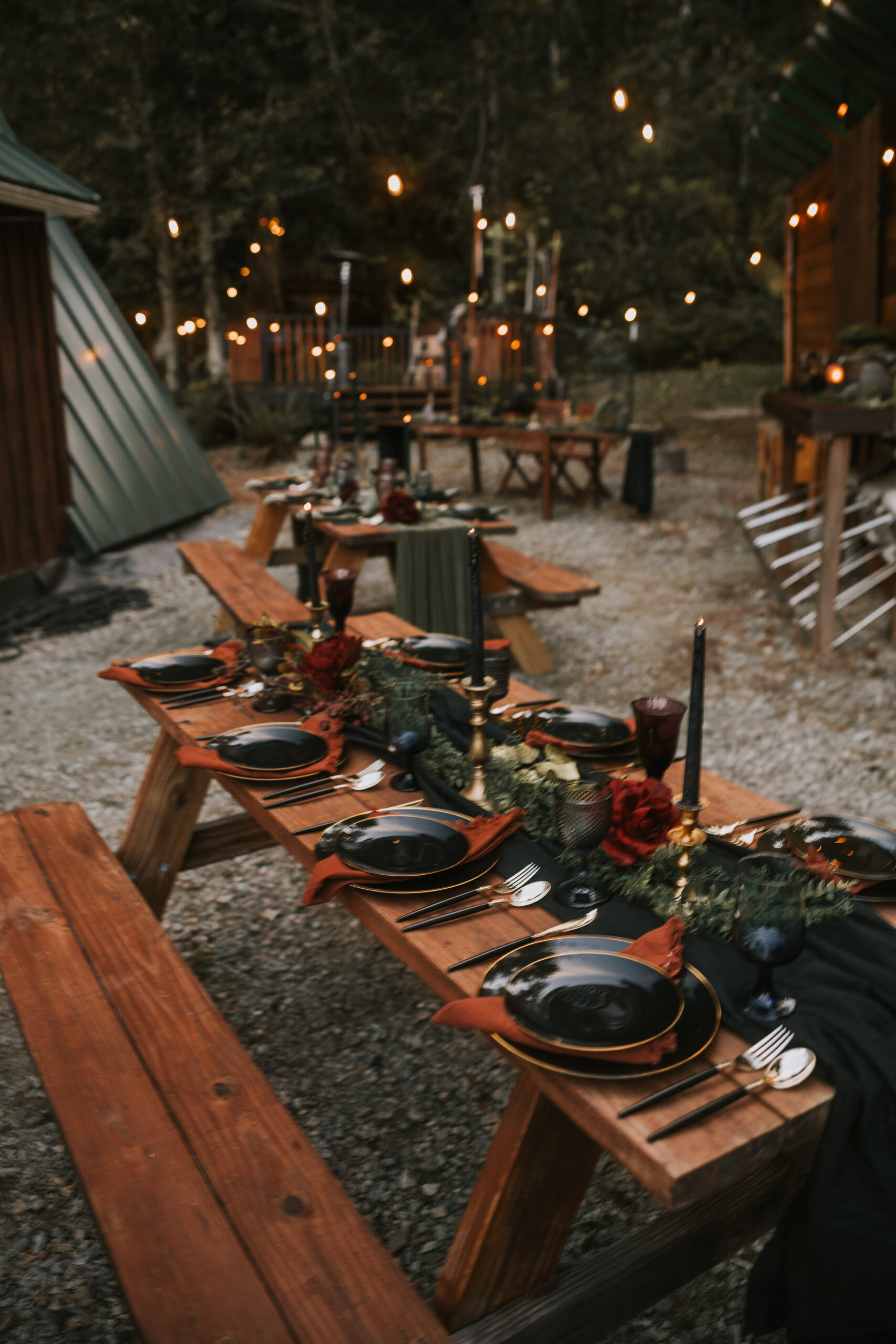 Long tables set with black and gold plates and rust napkins under string lights at dusk