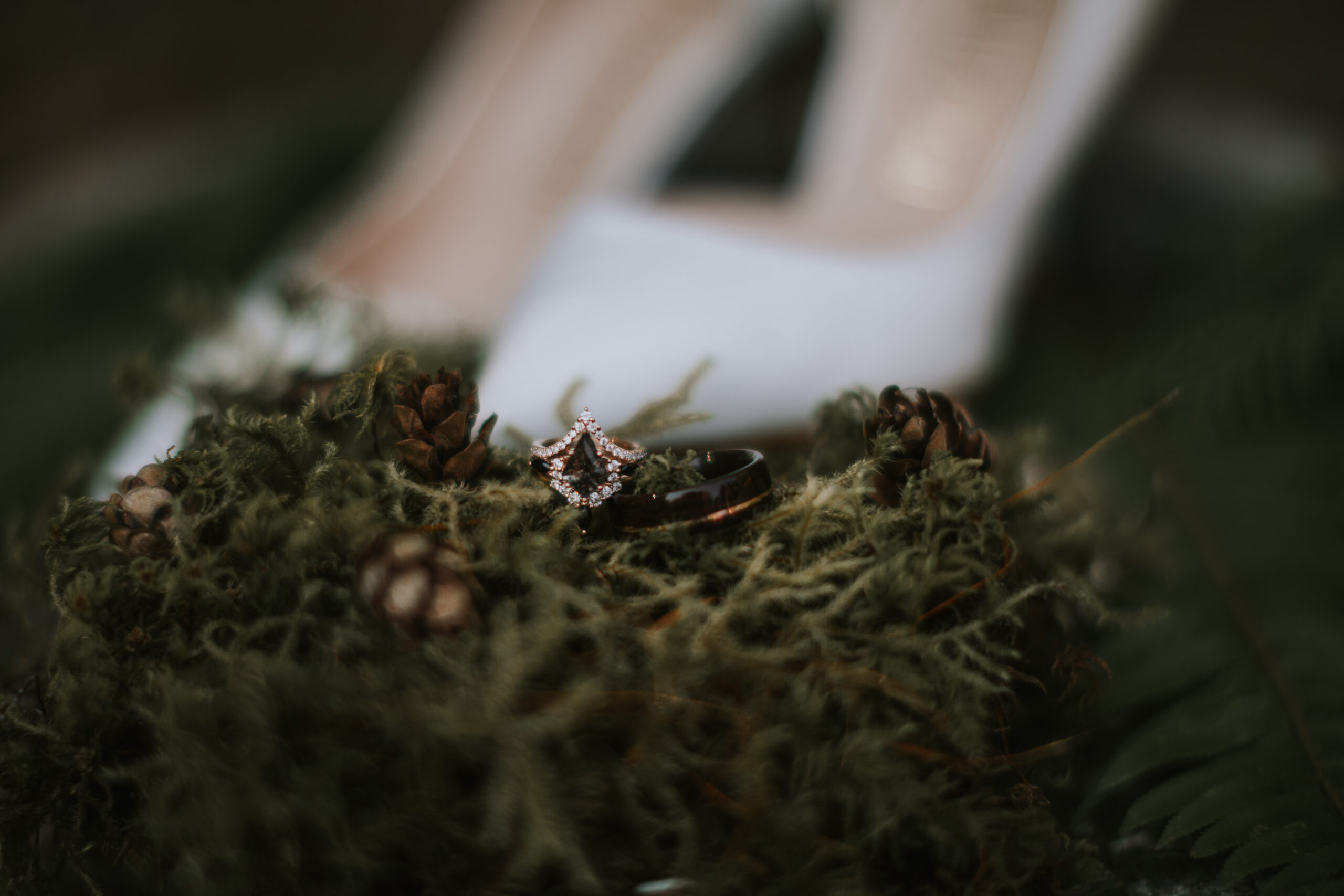 The engagement ring resting on moss and pinecones with the bride's shoes behind