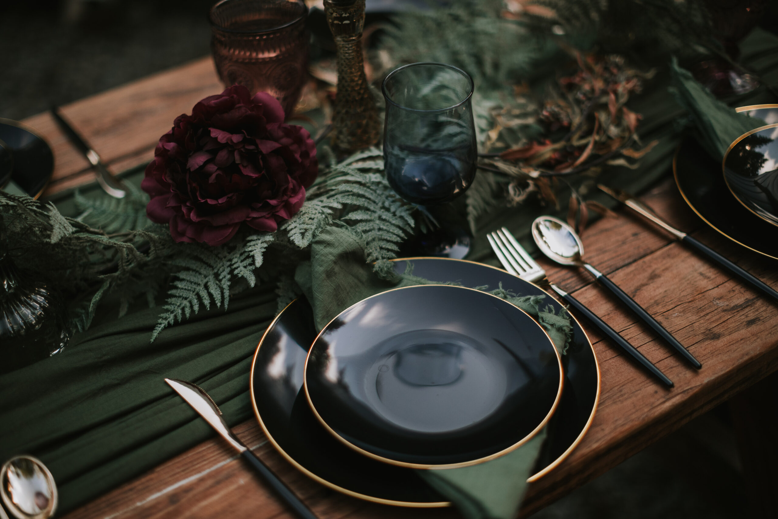 Place setting with dark gold rimmed plates, a burgundy bloom and fern along the runner