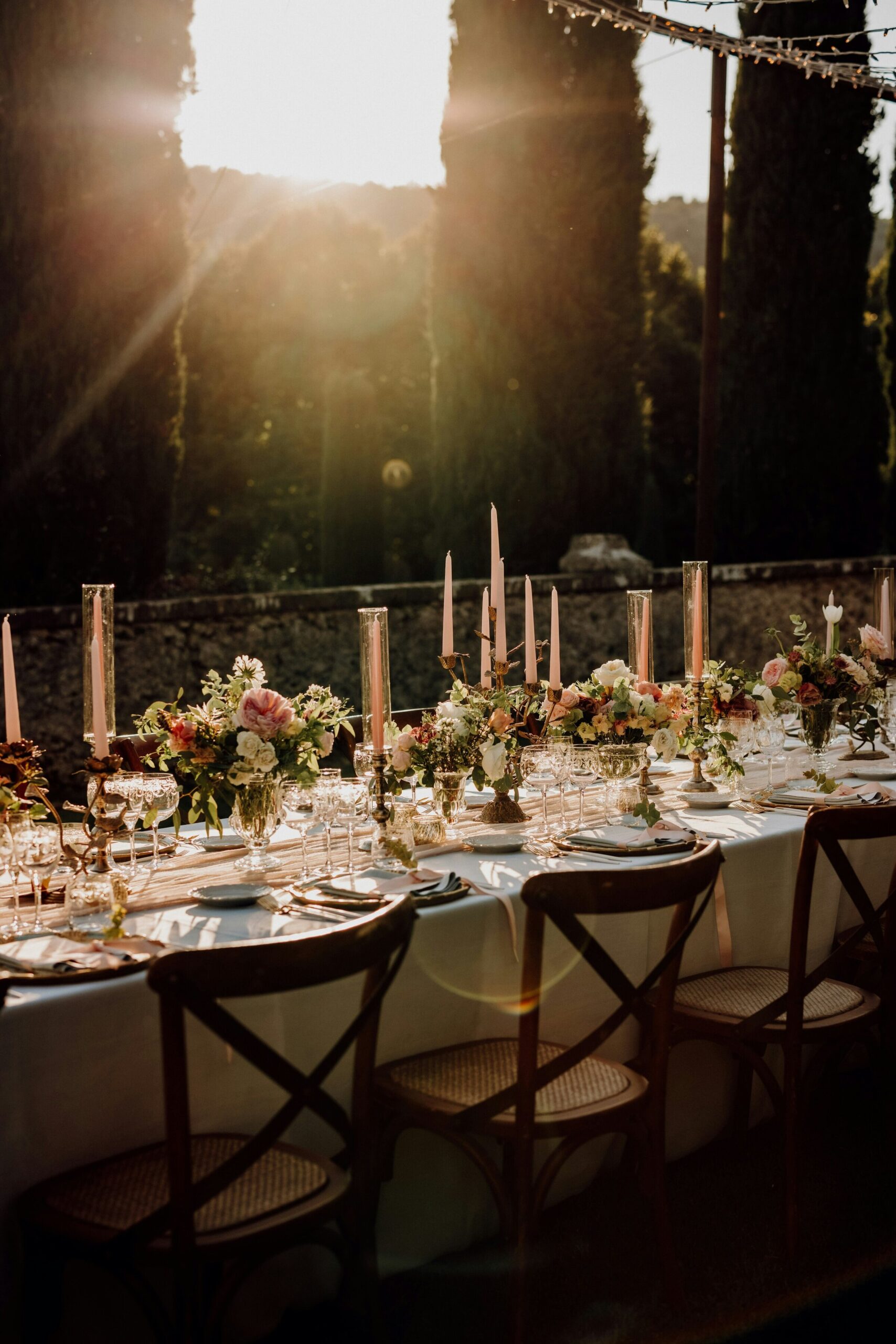 A long candlelit table at golden hour set with tapers in brass holders, glassware and low floral arrangements