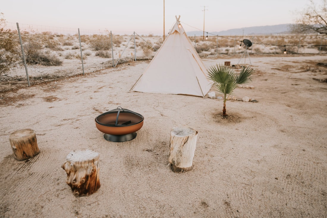 A fire pit ringed with stump seats and a canvas tipi on open desert at That Desert Space