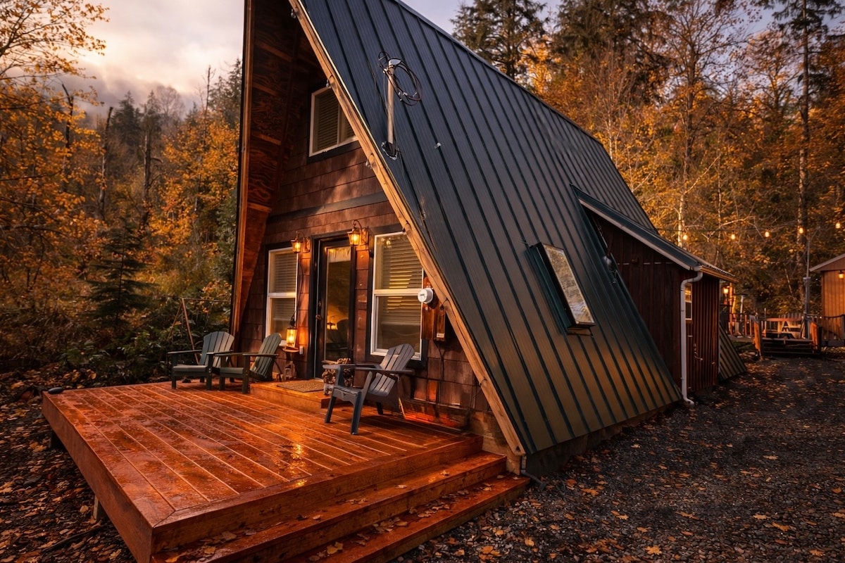 The A-frame from the side in autumn, cedar shingles and green metal roof with chairs on the wraparound deck